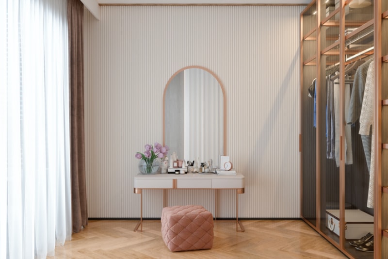 un placard ouvert à droite et des baies vitrées avec des rideaux blancs à gauche autour d'une coiffeuse blanche et d'un miroir au centre