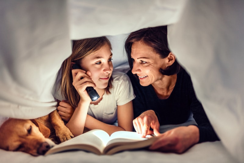 Mère, fille et chien allongés sur le lit sous une couverture tenant une lampe de poche et lisant un livre tard dans la nuit.