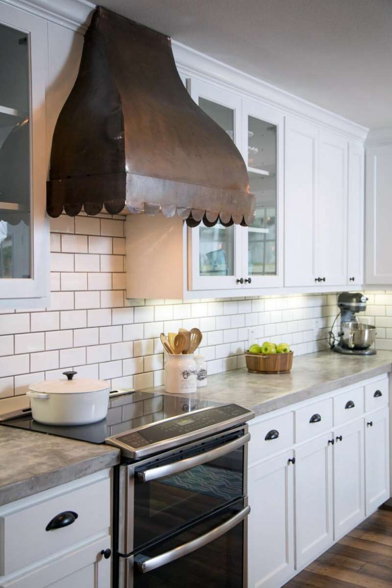 Une hotte de cuisinière rustique avec une bordure festonnée dans une cuisine avec des armoires blanches et un dosseret en carreaux blancs.