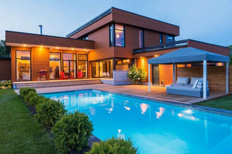 Une maison rectangulaire moderne avec une piscine éclairée située à côté d'un auvent de terrasse.