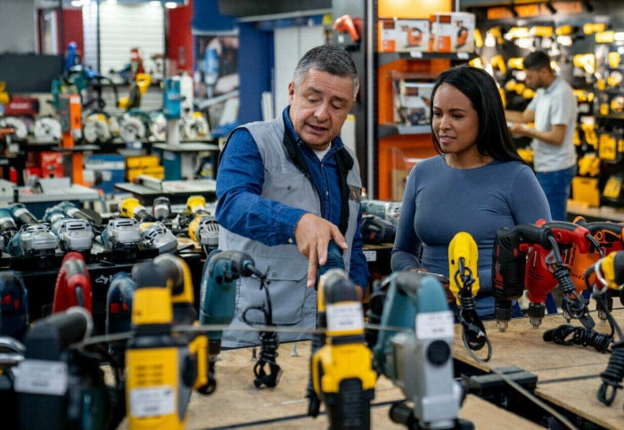 Un employé d'une quincaillerie parle à une femme des options de perceuse électrique sur une table.