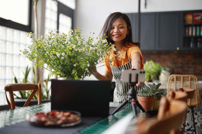 Une femme fleuriste vlogue sur son smartphone et son ordinateur portable, créant un tutoriel en ligne sur la composition d'un bouquet de fleurs à la maison.