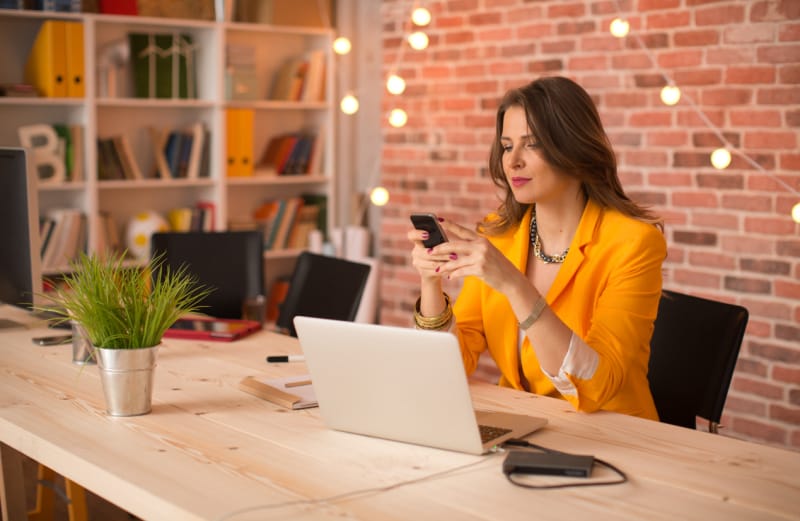 Une guirlande lumineuse est suspendue à un mur de briques derrière une femme assise à un bureau et regardant son téléphone.