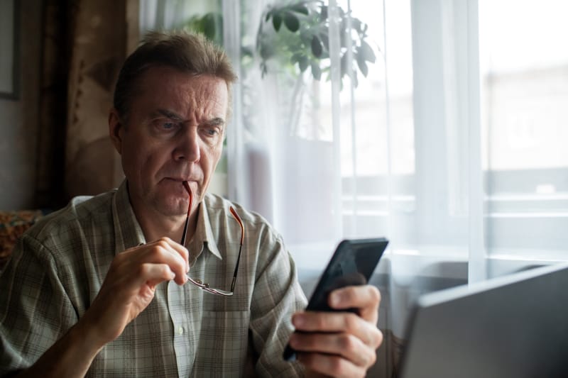 Un homme âgé à l'air inquiet regarde sérieusement son smartphone tout en étant assis devant un ordinateur portable.