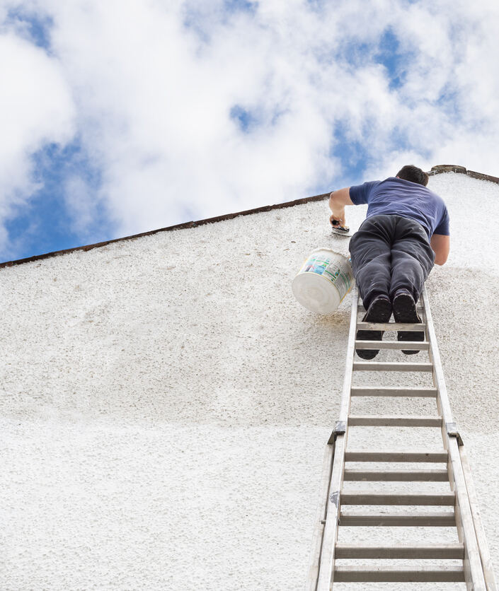 Photo de haute qualité d'un homme perché sur une échelle peignant un mur en crépi blanc sur le pignon d'une maison.