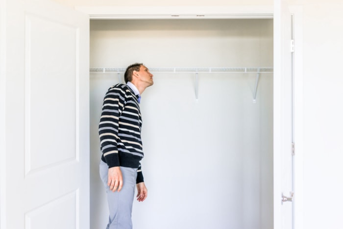 un homme en pull rayé et pantalon gris regarde dans le placard d'une chambre vide contre un mur blanc