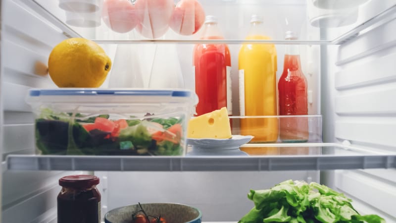 Vue rapprochée d'une étagère de réfrigérateur avec une salade à l'intérieur d'un Tupperware, un morceau de fromage non emballé et des bouteilles de jus et de condiments à l'arrière.