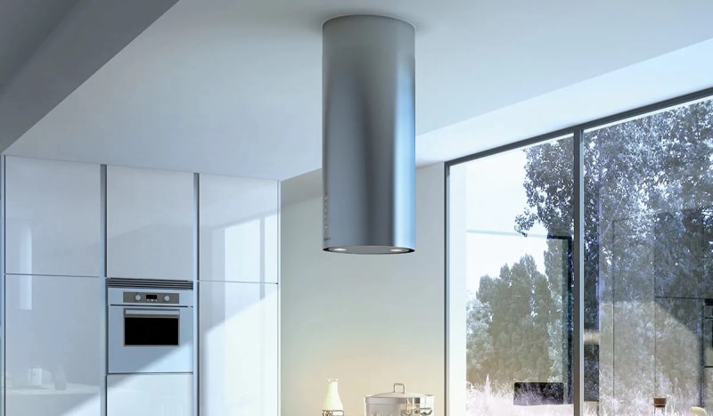 Une hotte de cuisinière cylindrique en acier inoxydable dans une cuisine blanche avec une grande fenêtre.