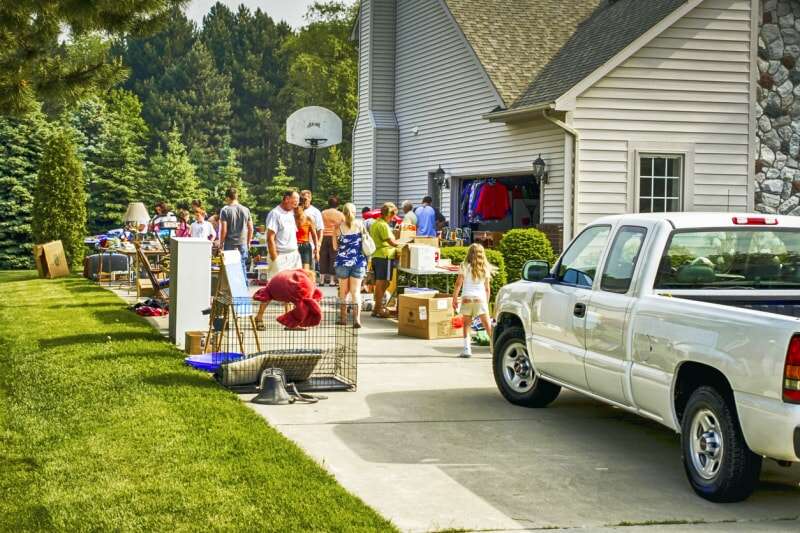 allée avec camionnette garée et tables installées pour la vente de garage et groupes de personnes faisant leurs courses à l'extérieur d'une grande maison