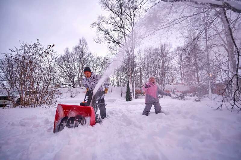 Un homme en veste blanche et chaud utilise une souffleuse à neige rouge tandis qu'un enfant en veste rose joue à proximité.