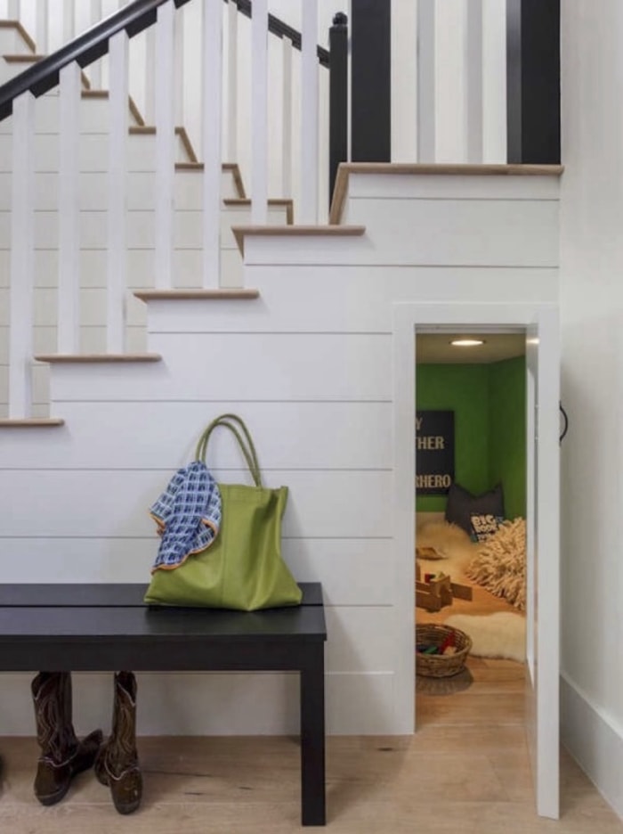 vue d'un lambris blanc sous un escalier avec des bottes de cow-boy sous un banc et une petite porte ouverte vers un vide sanitaire rempli de coussins, de tapis et de jouets