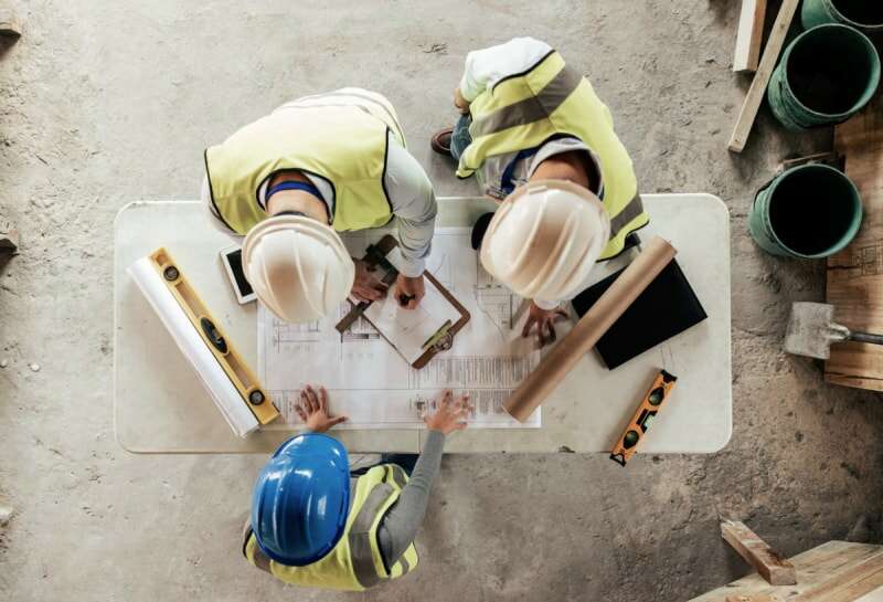 Vue aérienne de trois ouvriers portant des casques de protection et consultant un plan de construction dans une zone en cours de construction.