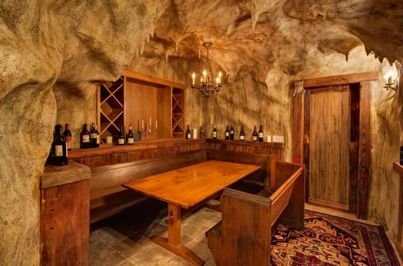 Une cave à vin avec une porte en bois, un sol en dalles de pierre, un tapis, une table et un banc en bois se trouve à l'intérieur des murs de la grotte.