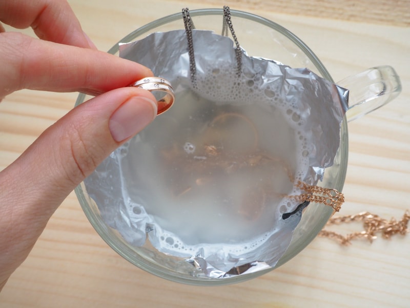 La main d'une femme place une bague dans une petite tasse à thé en verre contenant de l'eau avec d'autres bijoux en cours de nettoyage.