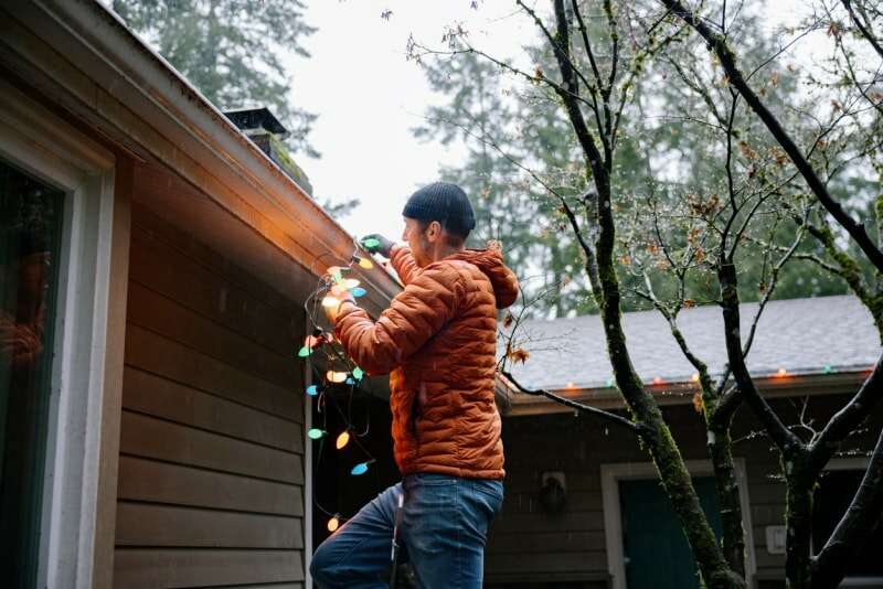 Un homme portant un manteau orange accroche des lumières de Noël sur sa maison par temps nuageux.