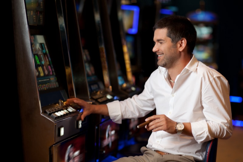 Un homme dans un casino est assis à une machine à sous, fumant un cigare et souriant.