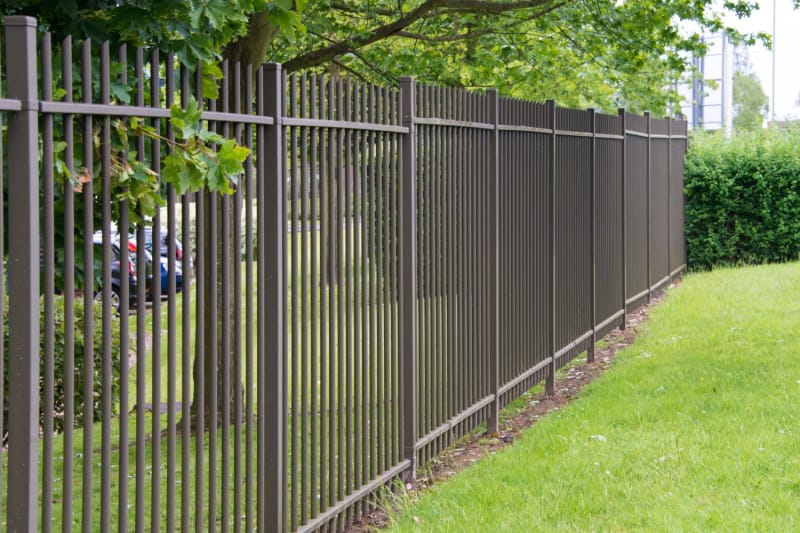 Une clôture en fer forgé noir divise une pelouse avec un parking entouré d'arbres.