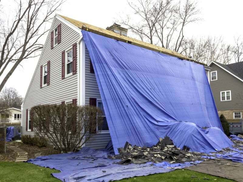 maison coloniale grise recouverte d'une grande bâche bleue avec des tuiles sur la pelouse pendant que le toit est remplacé