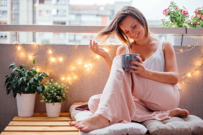 Une femme savoure son café sur un balcon avec des guirlandes lumineuses et des pots de fleurs.