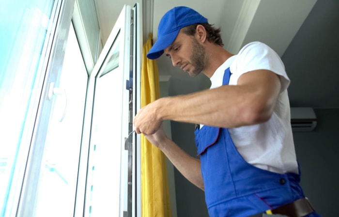 Un ouvrier vêtu d'un chapeau bleu et d'une salopette utilise un tournevis pour réparer le cadre d'une fenêtre dans une maison.