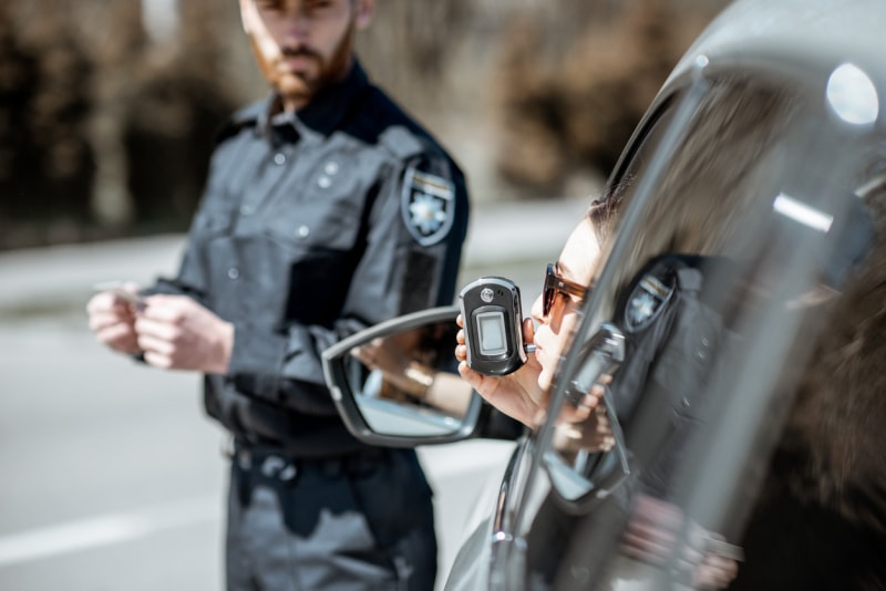 Vue de l'arrière du côté gauche d'une voiture noire alors que la conductrice passe l'alcootest en se penchant par la fenêtre et qu'un policier flou observe à distance.